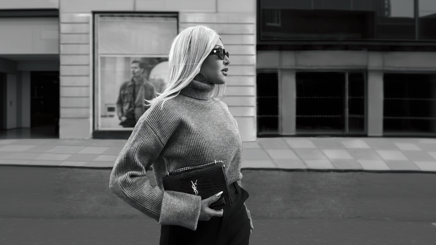 A black and white street-style photograph of a woman with platinum blonde hair wearing oversized sunglasses, a ribbed turtleneck sweater, and holding a black Saint Laurent clutch bag.